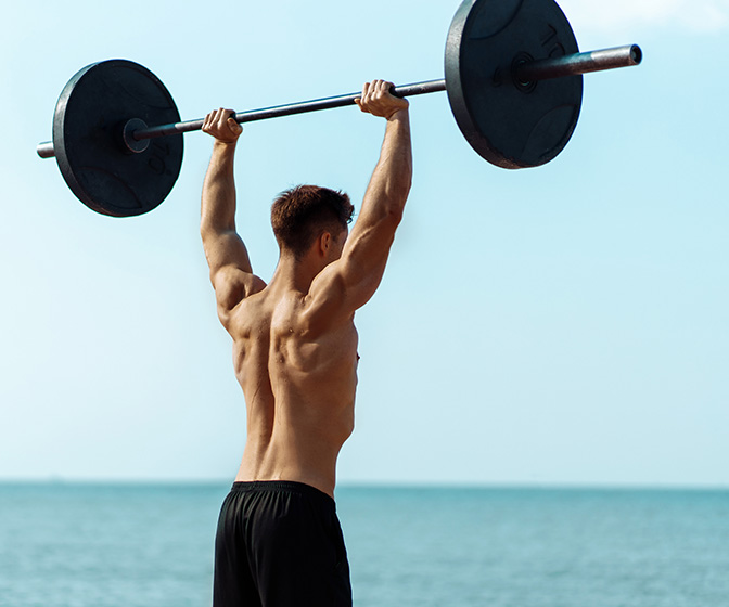 Muscular man training on the beach by the sea, doing exercises with a barbell for biceps, strong male press with a naked torso, sports concept